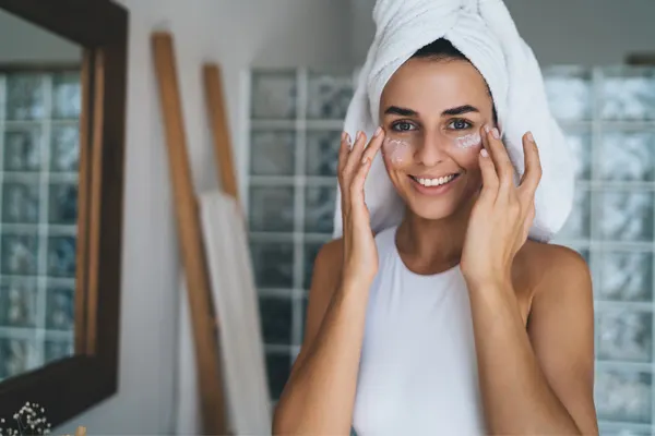 Roześmiana, młoda kobieta z białym ręcznikiem na głowie aplikuje opuszkami palców krem pod oczy .